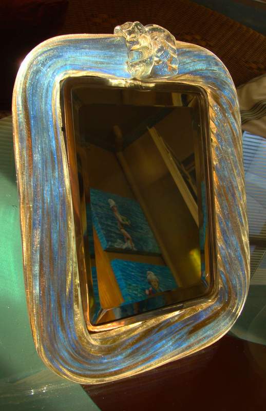 GlassFrameMirror1-sm.jpg :: This Murano twisted rope glass with gold leaf and silver dressing table mirror was made in Murano, Italy in the 1920's-30's. The mirror is almost magical in that the colour is an iridescent gold with shades of aqua. It changes with different light conditions.
The importance of this piece is reflected in its weight and construction. 
The mirror reclines at a 45 degree angle on a solid silver leg.  It is mounted on a similar frame of silver with a wooden back.  All appear to be original.  
Similar mirrors on the internet sell for $3000-$5000.  On sale at the Gallery for R14.500.
