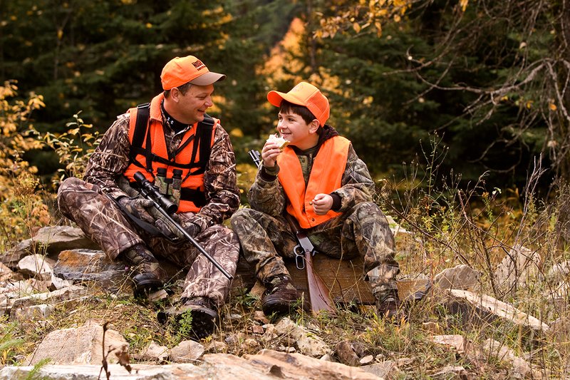 ©TC-Hunter Parent Child-D89995-00065.jpg :: Father wearing Advantage Max 4 camo w/florescent orange vest & hat hunts with his son wearing Realtree Brown camo and florescent orange vest/hat sitting together while on a hunt sharing conversation while eating lunch
