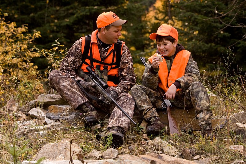 ©TC-Hunter Parent Child-D89995-00074.jpg :: Father wearing Advantage Max 4 camo w/florescent orange vest & hat hunts with his son wearing Realtree Brown camo and florescent orange vest/hat sitting together while on a hunt sharing conversation while eating lunch