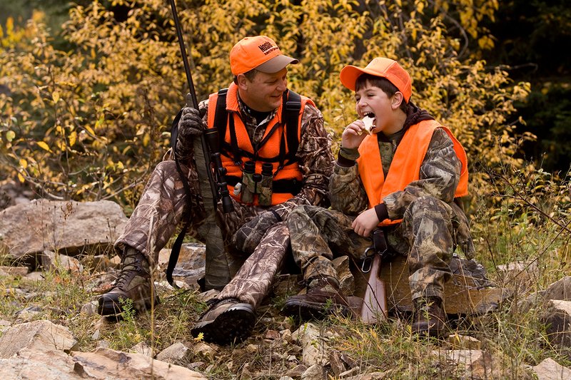 ©TC-Hunter Parent Child-D89995-00084.jpg :: Father wearing Advantage Max 4 camo w/florescent orange vest & hat hunts with his son wearing Realtree Brown camo and florescent orange vest/hat sitting together while on a hunt sharing conversation while eating lunch