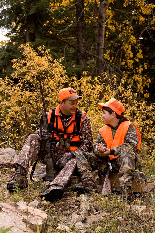 ©TC-Hunter Parent Child-D89995-00089.jpg :: Father wearing Advantage Max 4 camo w/florescent orange vest & hat hunts with his son wearing Realtree Brown camo and florescent orange vest/hat sitting together while on a hunt sharing conversation while eating lunch
