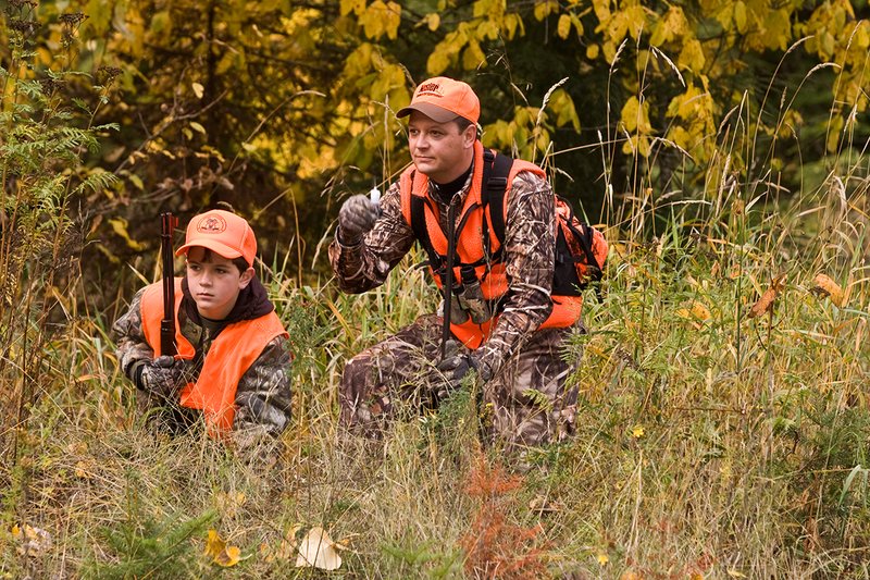 ©TC-Hunter Parent Child-D89995-00092.jpg :: Father wearing Advantage Max 4 camo w/florescent orange vest & hat uses a powder bottle to test the wind direction for his son wearing Realtree Brown camo and florescent orange vest/hat