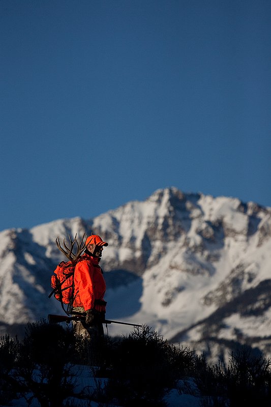©TC-Rifle Hunter in Snow-D089999-00591.jpg :: Rifle Hunter wearing florescent orange coat/hat pauses as he packs out the meat and rack of trophy mule deer buck with the grandeur of a snowcapped mountain peak in the background
