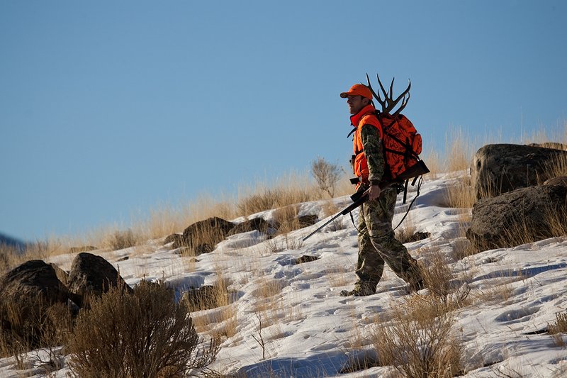 ©TC-Rifle Hunter in Snow-D089999-00651.jpg :: Rifle Hunter wearing Advantage Max1-HD camo with florescent orange hat/vest carrying a backpack with meat and rack of trophy mule deer along the crest of a mountain ridge