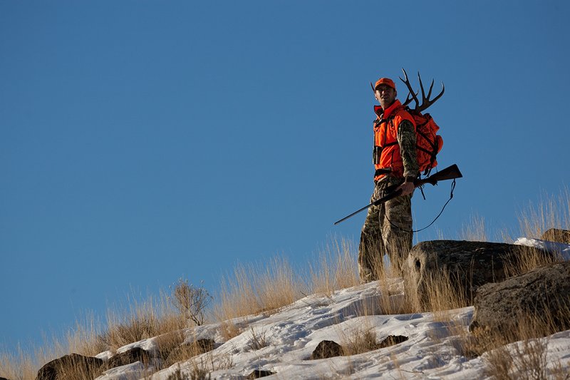 ©TC-Rifle Hunter in Snow-D089999-00659.jpg :: Rifle Hunter wearing Advantage Max1-HD camo with florescent orange hat/vest carrying a backpack with meat and rack of trophy mule deer along the crest of a mountain ridge