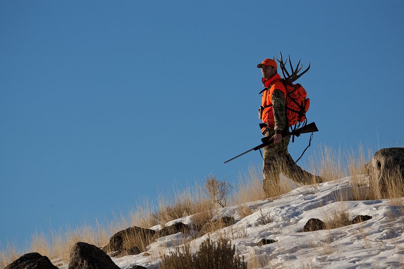 ©TC-Rifle Hunter in Snow-D089999-00663.jpg :: Rifle Hunter wearing Advantage Max1-HD camo with florescent orange hat/vest carrying a backpack with meat and rack of trophy mule deer along the crest of a mountain ridge