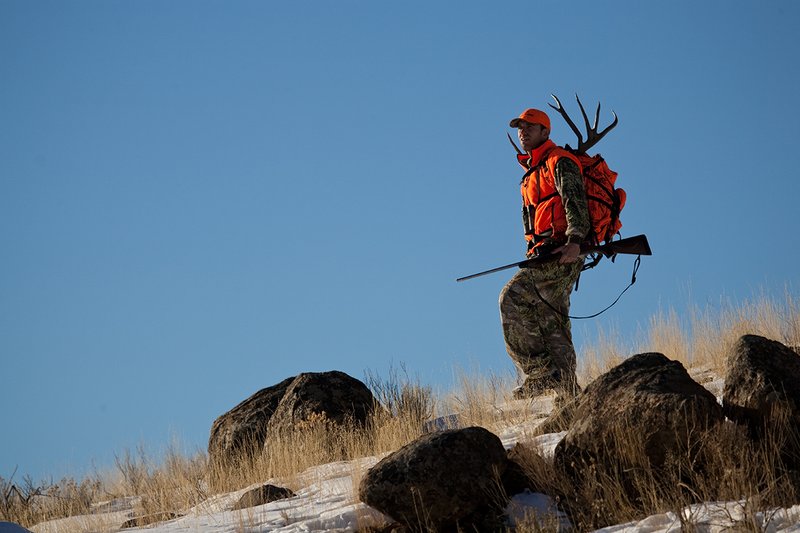 ©TC-Rifle Hunter in Snow-D089999-00672.jpg :: Rifle Hunter wearing Advantage Max1-HD camo with florescent orange hat/vest carrying a backpack with meat and rack of trophy mule deer along the crest of a mountain ridge