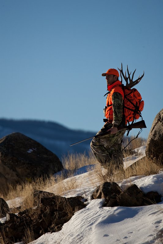 ©TC-Rifle Hunter in Snow-D089999-00678.jpg :: Rifle Hunter wearing Advantage Max1-HD camo with florescent orange hat/vest carrying a backpack with meat and rack of trophy mule deer along the crest of a mountain ridge