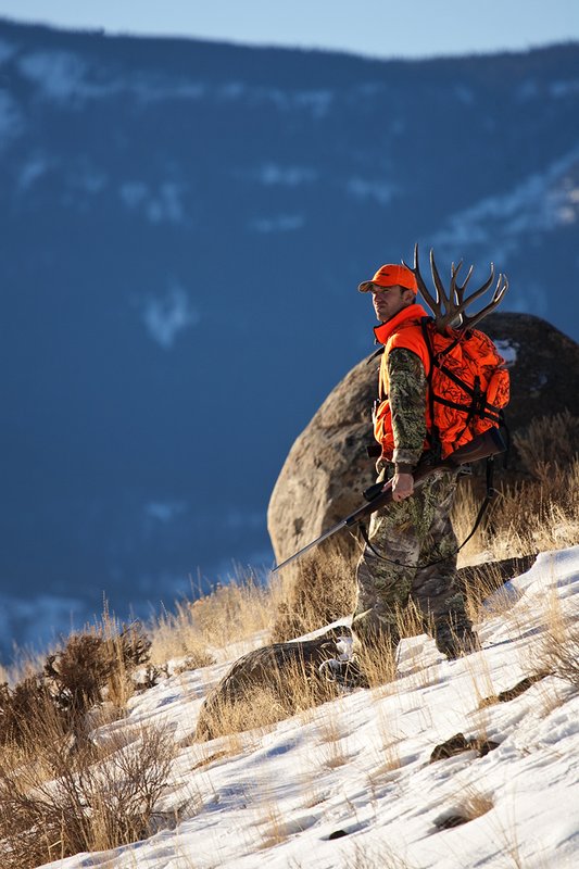 ©TC-Rifle Hunter in Snow-D089999-00687.jpg :: Rifle Hunter wearing Advantage Max1-HD camo with florescent orange hat/vest carrying a backpack with meat and rack of trophy mule deer off steep mountain ridge