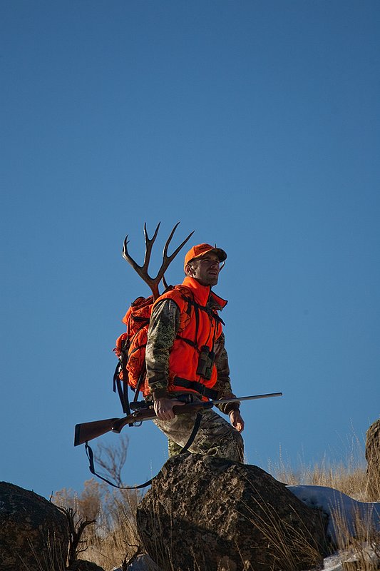 ©TC-Rifle Hunter in Snow-D089999-00690.jpg :: Rifle Hunter wearing Advantage Max1-HD camo with florescent orange hat/vest carrying a backpack with meat and rack of trophy mule deer along the crest of a mountain ridge