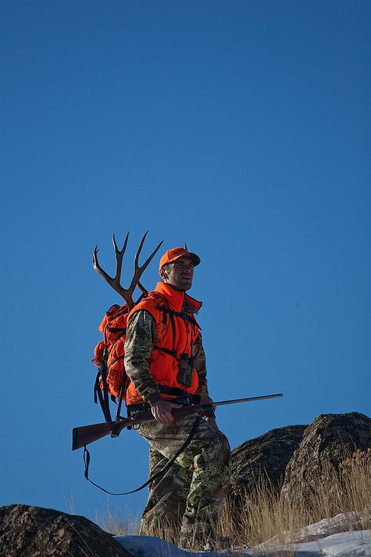 ©TC-Rifle Hunter in Snow-D089999-00693.jpg :: Rifle Hunter wearing Advantage Max1-HD camo with florescent orange hat/vest carrying a backpack with meat and rack of trophy mule deer along the crest of a mountain ridge