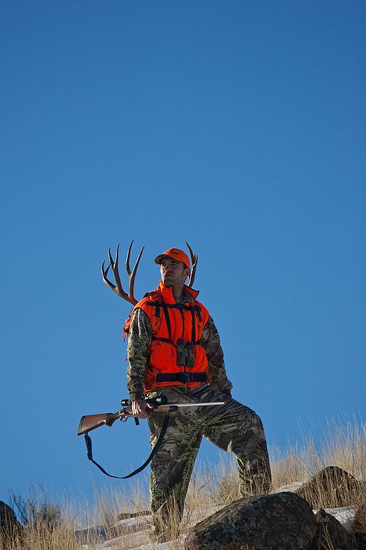 ©TC-Rifle Hunter in Snow-D089999-00699.jpg :: Rifle Hunter wearing Advantage Max1-HD camo with florescent orange hat/vest carrying a backpack with meat and rack of trophy mule deer along the crest of a mountain ridge