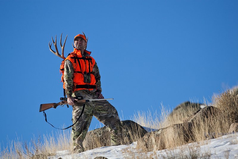 ©TC-Rifle Hunter in Snow-D089999-00708.jpg :: Rifle Hunter wearing Advantage Max1-HD camo with florescent orange hat/vest carrying a backpack with meat and rack of trophy mule deer along the crest of a mountain ridge