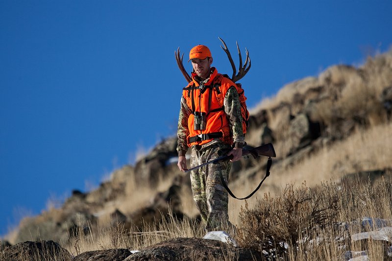 ©TC-Rifle Hunter in Snow-D089999-00711.jpg :: Rifle Hunter wearing Advantage Max1-HD camo with florescent orange hat/vest carrying a backpack with meat and rack of trophy mule deer along the crest of a mountain ridge