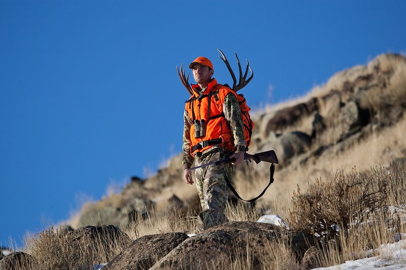 ©TC-Rifle Hunter in Snow-D089999-00713.jpg :: Rifle Hunter wearing Advantage Max1-HD camo with florescent orange hat/vest carrying a backpack with meat and rack of trophy mule deer along the crest of a mountain ridge
