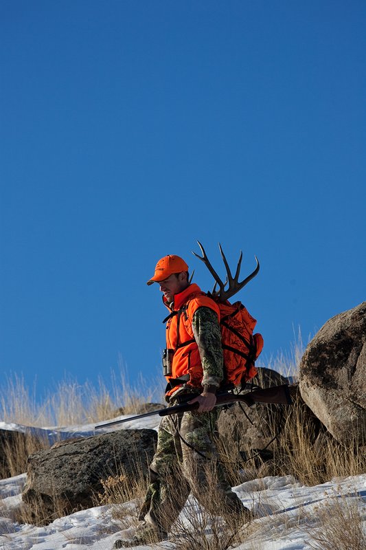 ©TC-Rifle Hunter in Snow-D089999-00716.jpg :: Rifle Hunter wearing Advantage Max1-HD camo with florescent orange hat/vest carrying a backpack with meat and rack of trophy mule deer along the crest of a mountain ridge