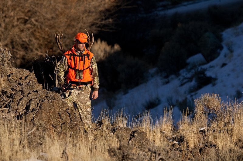 ©TC-Rifle Hunter in Snow-D089999-00819.jpg :: Rifle Hunter wearing Advantage Max1-HD camo with florescent orange hat/vest carrying a backpack with meat and rack of trophy mule deer along tall sage and rocky hillside