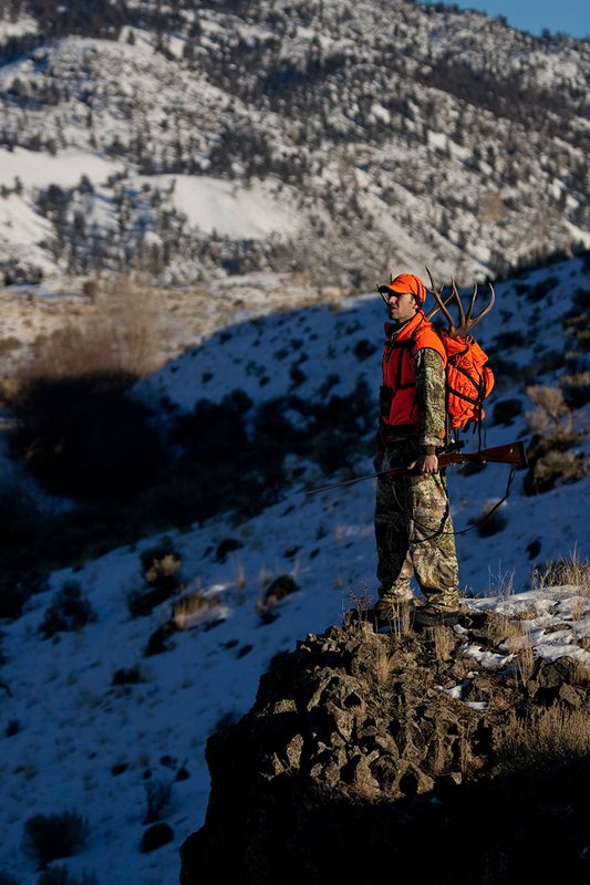 ©TC-Rifle Hunter in Snow-D089999-00838.jpg :: Rifle Hunter wearing Advantage Max1-HD camo with florescent orange hat/vest carrying a backpack with meat and rack of trophy mule deer on a rocky sage covered ridge