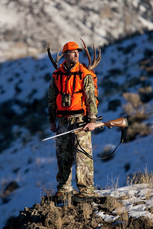 ©TC-Rifle Hunter in Snow-D089999-00840.jpg :: Rifle Hunter wearing Advantage Max1-HD camo with florescent orange hat/vest carrying a backpack with meat and rack of trophy mule deer on a rocky sage covered ridge