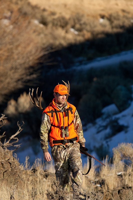 ©TC-Rifle Hunter in Snow-D089999-00843.jpg :: Rifle Hunter wearing Advantage Max1-HD camo with florescent orange hat/vest carrying a backpack with meat and rack of trophy mule deer on a rocky sage covered ridge