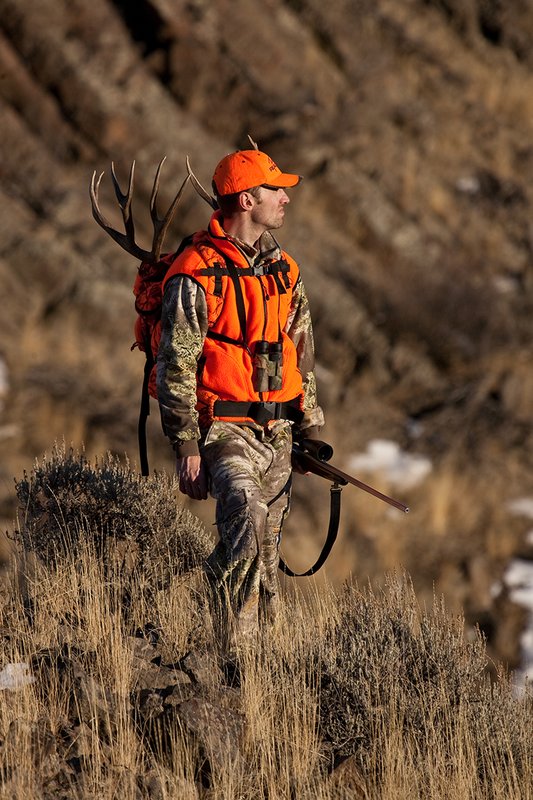 ©TC-Rifle Hunter in Snow-D089999-00851.jpg :: Rifle Hunter wearing Advantage Max1-HD camo with florescent orange hat/vest carrying a backpack with meat and rack of trophy mule deer