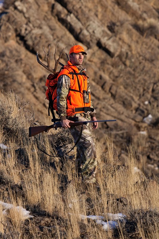 ©TC-Rifle Hunter in Snow-D089999-00864.jpg :: Rifle Hunter wearing Advantage Max1-HD camo with florescent orange hat/vest carrying a backpack with meat and rack of trophy mule deer
