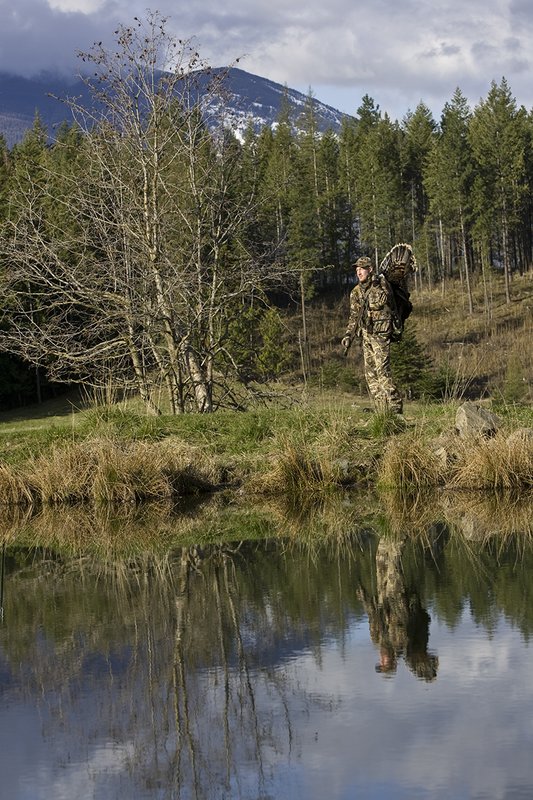 ©TC-Turkey Hunting-D90006-00405.jpg :: Turkey Hunter wearing Advantage Max 4HD Camo carrying Merriam Wild turkey out of the woods. Image of hunter and mountains reflected on surface of the pond.