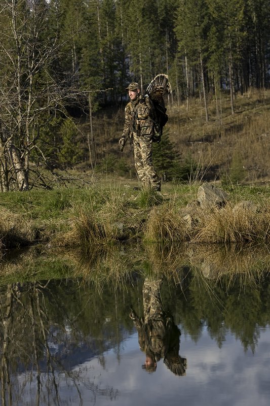 ©TC-Turkey Hunting-D90006-00411.jpg :: Turkey Hunter wearing Advantage Max 4HD Camo carrying Merriam Wild turkey out of the woods. Image of hunter and mountains reflected on surface of the pond.