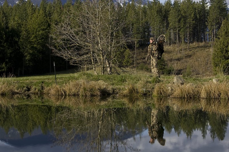 ©TC-Turkey Hunting-D90006-00414.jpg :: Turkey Hunter wearing Advantage Max 4HD Camo carrying Merriam Wild turkey out of the woods. Image of hunter and mountains reflected on surface of the pond.