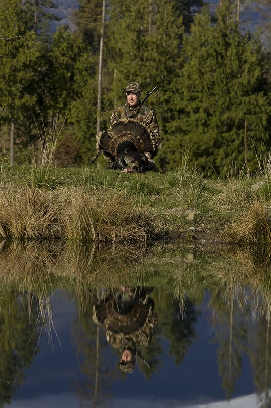 ©TC-Turkey Hunting-D90006-00417.jpg :: Turkey Hunter wearing Advantage Max 4HD Camo admiring his Merriam Long bearded gobbler at edge of pond.  Image of hunter and mountains reflected on surface of the pond.