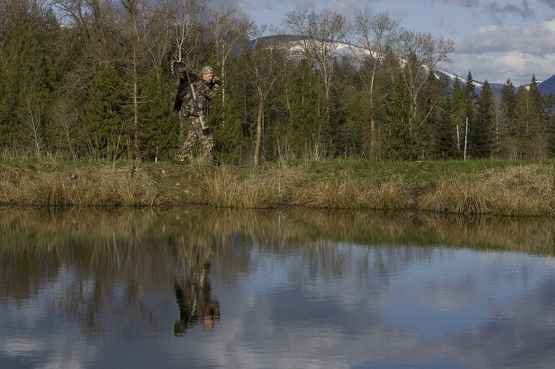 ©TC-Turkey Hunting-D90006-00435.jpg :: Turkey Hunter wearing Advantage Max 4HD Camo carrying Merriam Wild turkey out of the woods. Image of hunter and mountains reflected on surface of the pond.
