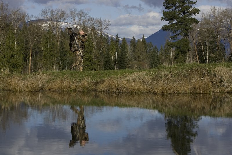 ©TC-Turkey Hunting-D90006-00438.jpg :: Turkey Hunter wearing Advantage Max 4HD Camo carrying Merriam Wild turkey out of the woods. Image of hunter and mountains reflected on surface of the pond.
