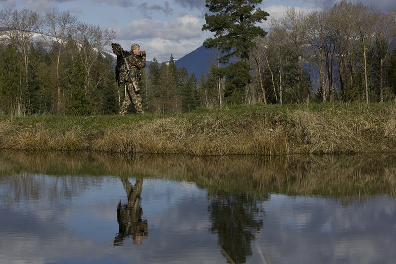 ©TC-Turkey Hunting-D90006-00441.jpg :: Turkey Hunter wearing Advantage Max 4HD Camo carrying Merriam Wild turkey out of the woods. Image of hunter and mountains reflected on surface of the pond.