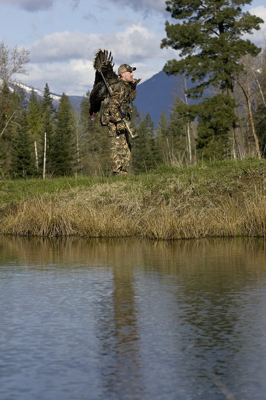 ©TC-Turkey Hunting-D90006-00449.jpg :: Turkey Hunter wearing Advantage Max 4HD Camo carrying Merriam Wild turkey out of the woods. Image of hunter and mountains reflected on surface of the pond.