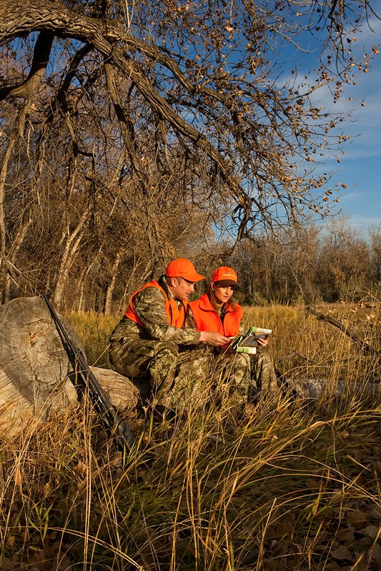 TC-Hunt-Women Big Game-D89992-00306.jpg :: Woman and man rifle hunting, river bottom, cottonwood trees, Max 1 camo, orange vest, hat, Elite Bushnell optics, looking at map, compass, naviagting,
