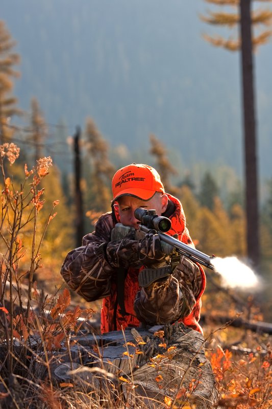 TC-Muzzleloader Hunters-D90008-00333.jpg :: Muzzleloader hunter wearing Advantage Max4-HD camo and Realtree Orange Vest shooting CVA Muzzleloader topped with Leuopold Omega Scope with smoke from muzzleoader coming from barrel, Larch trees in fall colors backlit in background