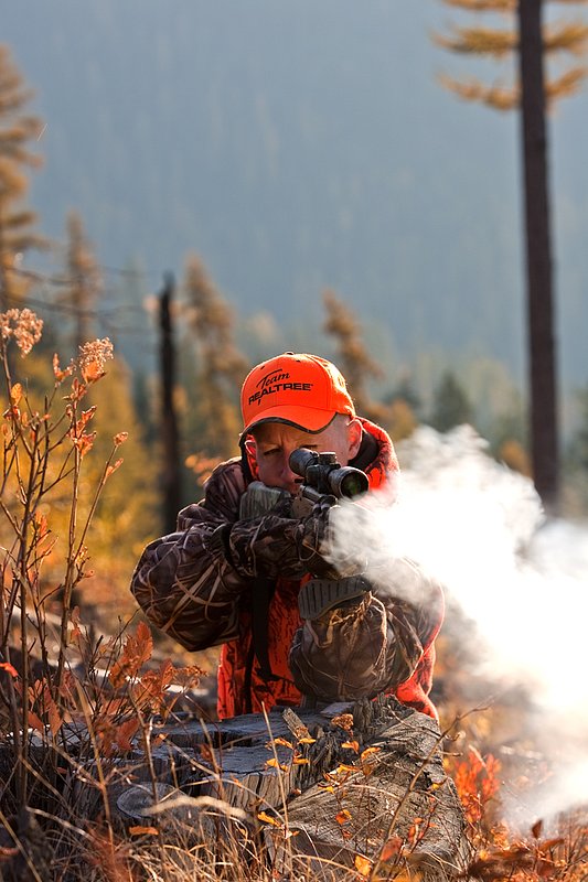 TC-Muzzleloader Hunters-D90008-00334.jpg :: Muzzleloader hunter wearing Advantage Max4-HD camo and Realtree Orange Vest shooting CVA Muzzleloader topped with Leuopold Omega Scope with smoke from muzzleoader coming from barrel, Larch trees in fall colors backlit in background