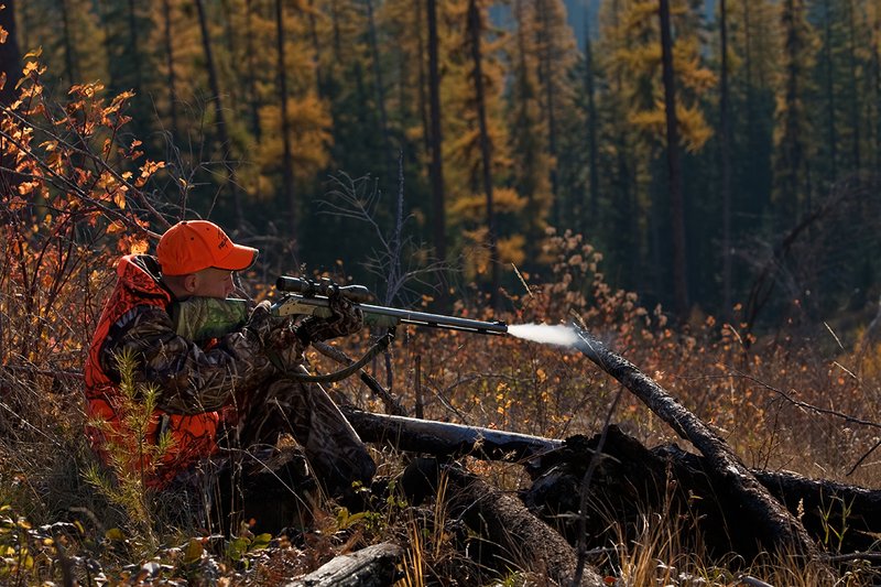 TC-Muzzleloader Hunters-D90008-00338.jpg :: Muzzleloader hunter wearing Advantage Max4-HD camo and Realtree Orange Vest shooting CVA Muzzleloader topped with Leuopold Omega Scope with smoke from muzzleoader coming from barrel, Larch trees in fall colors backlit in background