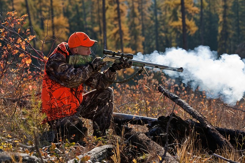 TC-Muzzleloader Hunters-D90008-00354.jpg :: Muzzleloader hunter wearing Advantage Max4-HD camo and Realtree Orange Vest shooting CVA Muzzleloader topped with Leuopold Omega Scope with smoke from muzzleoader coming from barrel, Larch trees in fall colors backlit in background
