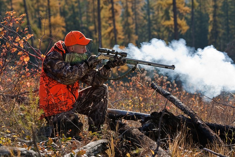 TC-Muzzleloader Hunters-D90008-00355.jpg :: Muzzleloader hunter wearing Advantage Max4-HD camo and Realtree Orange Vest shooting CVA Muzzleloader topped with Leuopold Omega Scope with smoke from muzzleoader coming from barrel, Larch trees in fall colors backlit in background