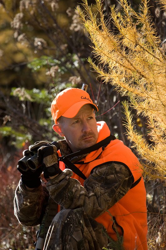 TC-Muzzleloader Hunters-D90008-00824.jpg :: Muzzleloader Hunter wearing Realtree AP Brown camo and a florescent orange vest and hat crouched down in a ground blind of pines and Western Larch glassing with Nikon 8x42 binocular for game