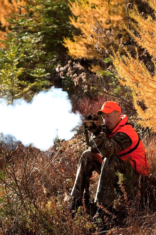 TC-Muzzleloader Hunters-D90008-00838.jpg :: Muzzleloader Hunter wearing Realtree AP Brown camo and a florescent orange vest and hat crouched down shooting his CVA muzzleloader amist pines and Western Larch