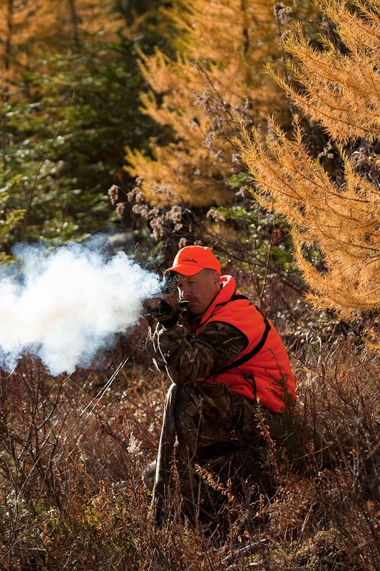 TC-Muzzleloader Hunters-D90008-00853.jpg :: Muzzleloader Hunter wearing Realtree AP Brown camo and a florescent orange vest and hat crouched down shooting his CVA muzzleloader amist pines and Western Larch