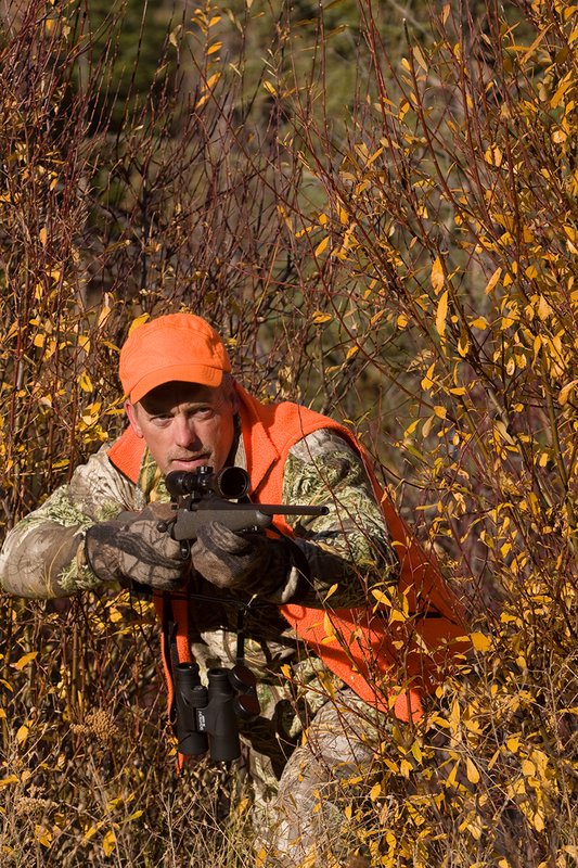 TC-Rifle Hunters-D90000-01349.jpg :: Rifle hunter wearing Realtree Max 1 camo and a florescent orange vest and hat stepping out from willow with a custom Remington Model 700 topped with a Sightron 3x9 Big Sky Rifle scope raised ready to shoot.