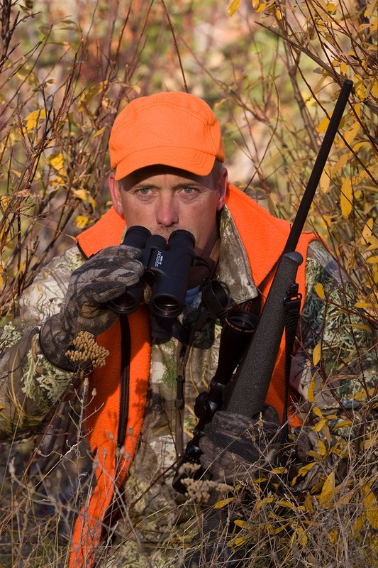 TC-Rifle Hunters-D90000-01352.jpg :: Rifle hunter wearing Realtree Max 1 camo and a florescent orange vest and hat crouched in willows watching game animals using a Sightron 10x42 binocular