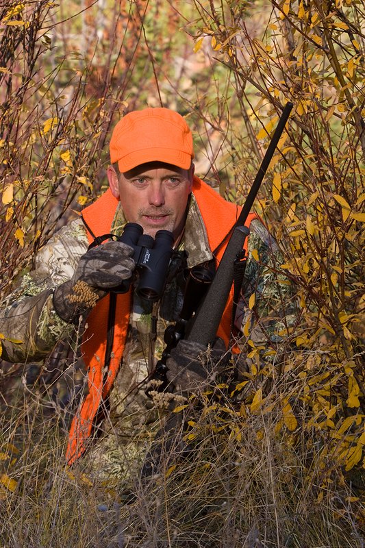 TC-Rifle Hunters-D90000-01353.jpg :: Rifle hunter wearing Realtree Max 1 camo and a florescent orange vest and hat crouched in willows watching game animals using a Sightron 10x42 binocular