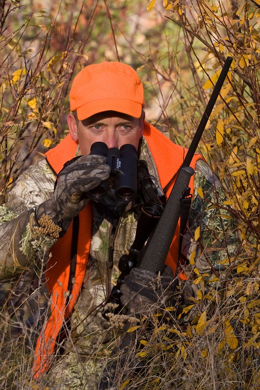 TC-Rifle Hunters-D90000-01355.jpg :: Rifle hunter wearing Realtree Max 1 camo and a florescent orange vest and hat crouched in willows watching game animals using a Sightron 10x42 binocular