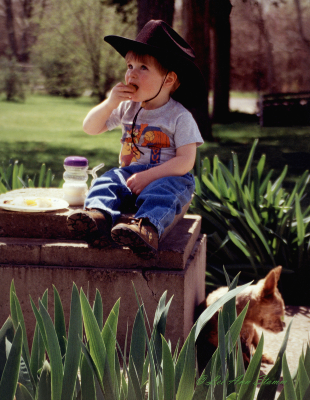 Little Cowboy Eats Lunch FINAL.jpg :: Little boy eats lunch on the lid of a cistern with a small dog by his side.  He is surrounded by iris leaves.  Taken in Lone Jack, Mo
