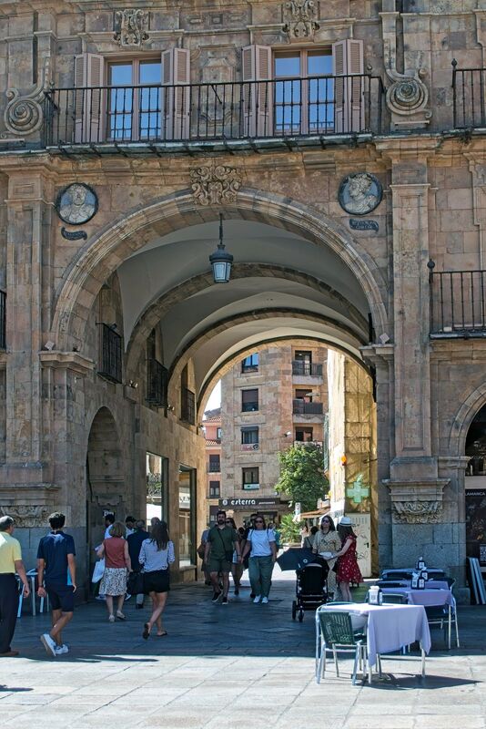 DC 22-0794-040.jpg :: Saga Douro River Cruise, Salamanca. Spain: The famous Plaza Mayor in Salamanca. Widely considered one of the most beautiful squares in the country, it is a significant example of Spanish Baroque architecture built in the 18th century. The Plaza Mayor is in the heart of Salamanca, a pedestrianized area that serves as the city's main social hub. It was primarily designed by architect Alberto Churriguera and completed in 1755. The buildings feature a uniform design of three stories, with arcades lining the perimeter and medallions depicting historic figures above the arches. Historically used for markets, bullfights, and public ceremonies, today the square is lined with numerous shops, restaurants, and cafes. It frequently hosts local events, concerts, and festivals, which explains the stage and seating setup. This archway is known as the \