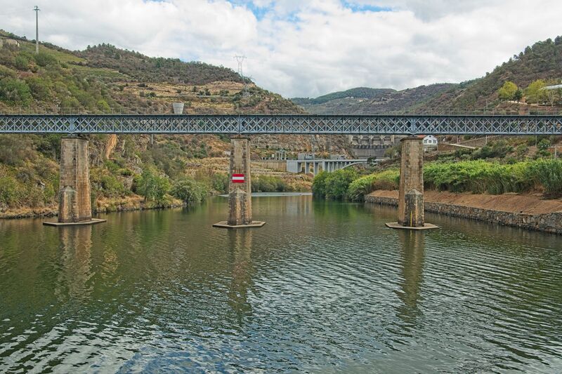 DC 22-0797-019.jpg :: Saga Douro River Cruise, Pocinho to Pinhao, River Douro: The Carrazeda de Ansiães railway bridge (also known as the Foz-Tua bridge) crossing the Douro River in Northern Portugal, a region known for its terraced vineyards. This striking iron truss bridge is located near the confluence of the Tua and Douro rivers in the municipality of Carrazeda de Ansiães. The bridge is situated in the heart of the Alto Douro region, which is a UNESCO World Heritage site known for its Port wine production. It is a railway bridge, part of the historical Douro railway line. The original structure was built in the late 19th or early 20th century, but a new bridge was constructed in 1980, featuring a mix of reinforced concrete for the access structures and a central metal truss. Photo taken 02/09/2022